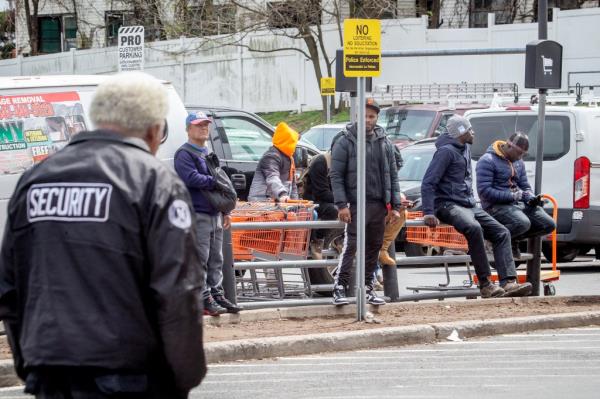 Security guard walking near migrants in the parking lot of a Home Depot store in Bronx, New York, assisting with loading purchases into vehicles